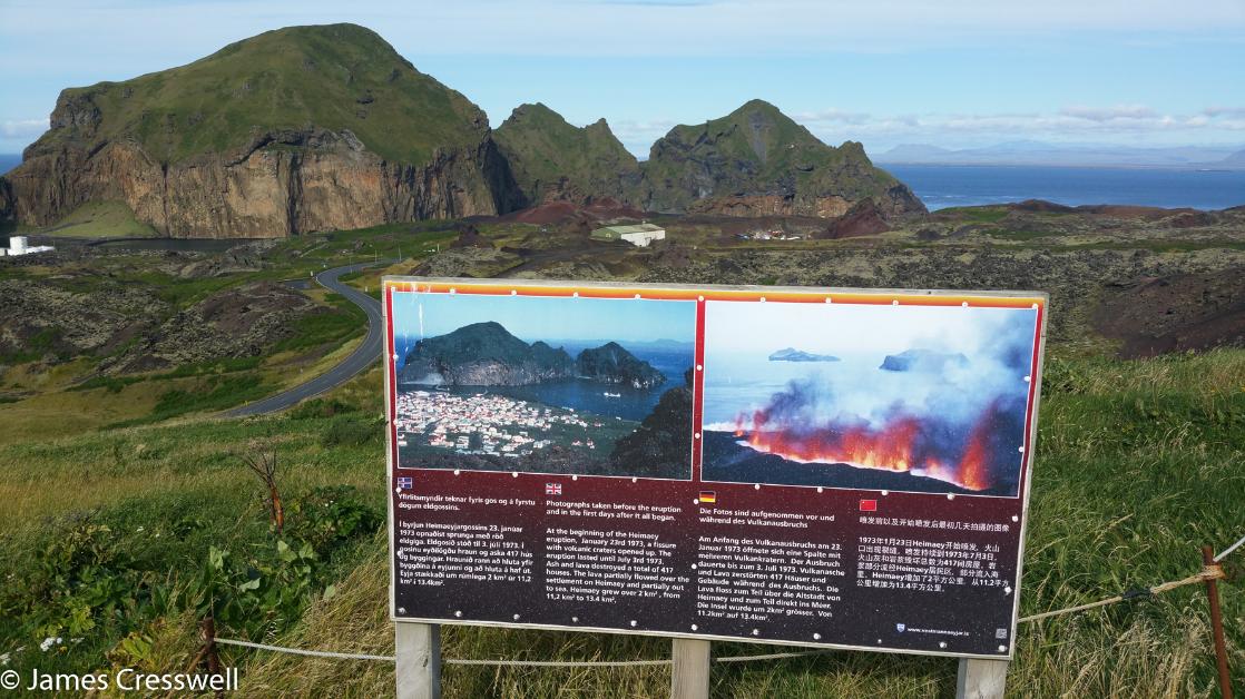 A photograph of a sign showing a volcano erupting in front of the location where the volcanic eruption occurred, at Vestmannaeyjar, taken on the GeoWorld Travel Iceland volcano trip and geology holiday
