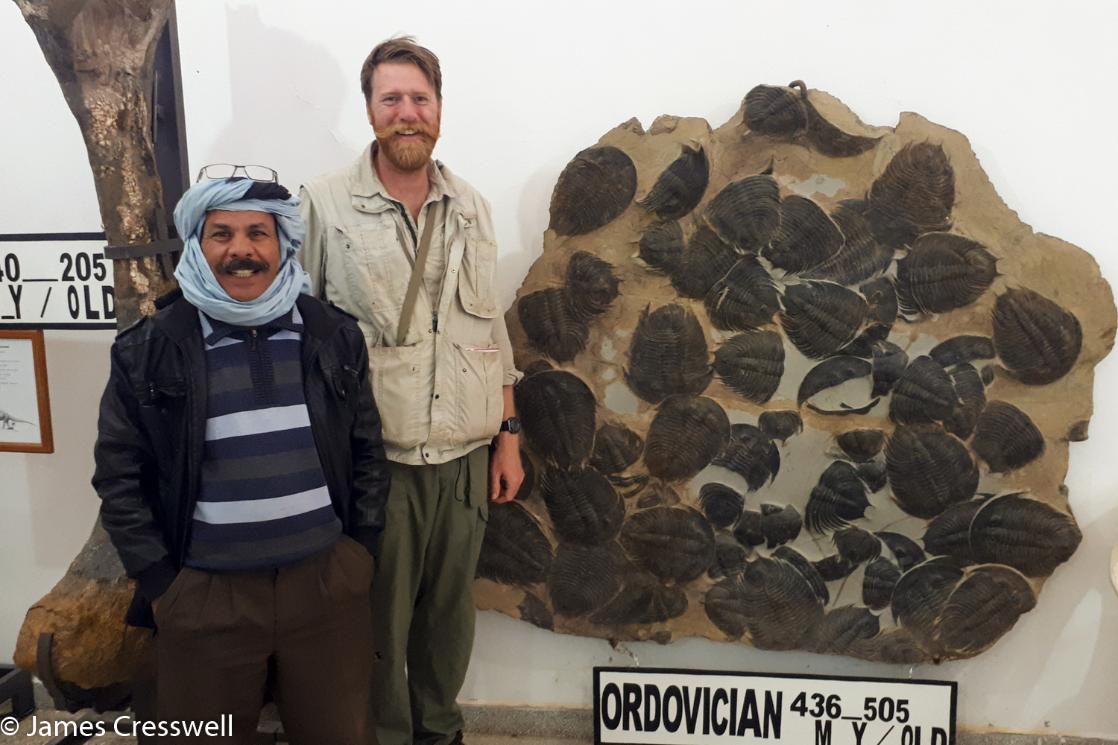A photograph of James Cresswell and the curator of the Erfoud Museum in front of giant trilobites, taken on a GeoWorld Travel geology and museum trip, tour and holiday