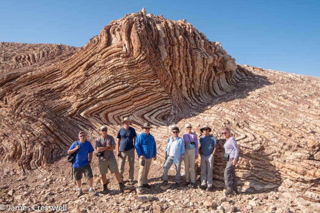 A photograph of eight people standing in front of red and white coloured folded rock, the Mother of All Outcrops in radiolarian chert, taken on a GeoWorld Travel Oman geology trip, tour and holiday