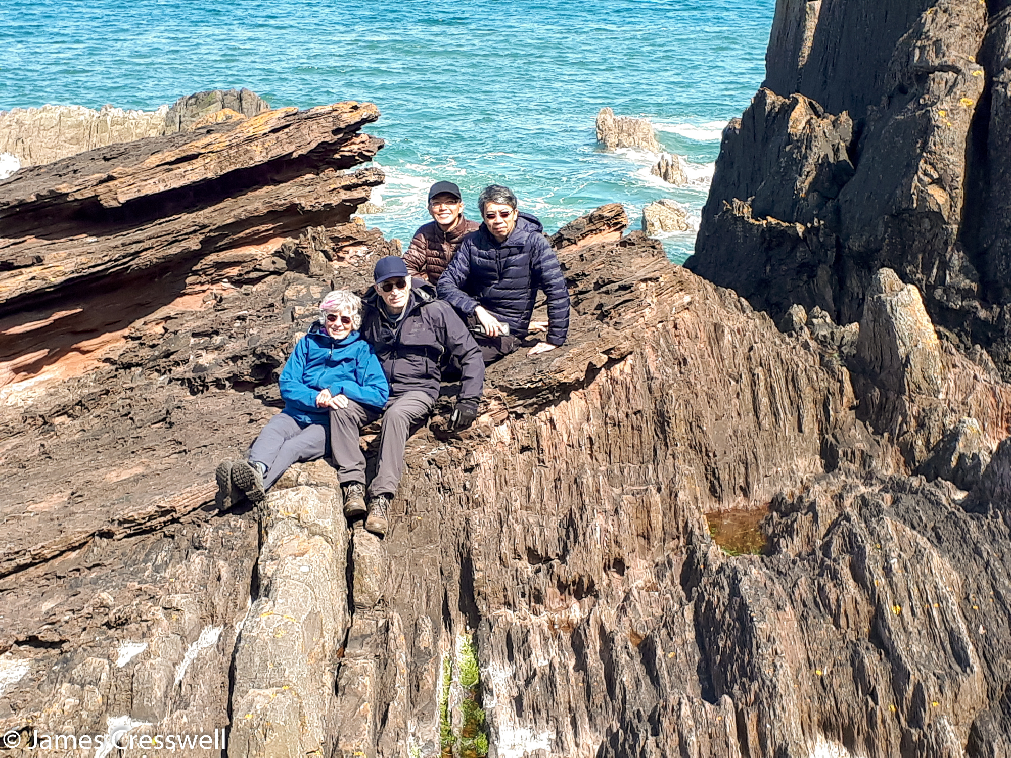A photograph of four people sitting on a geological formation, Siccar Point, taken on a GeoWorld Travel Scotland geology trip, tour and holiday