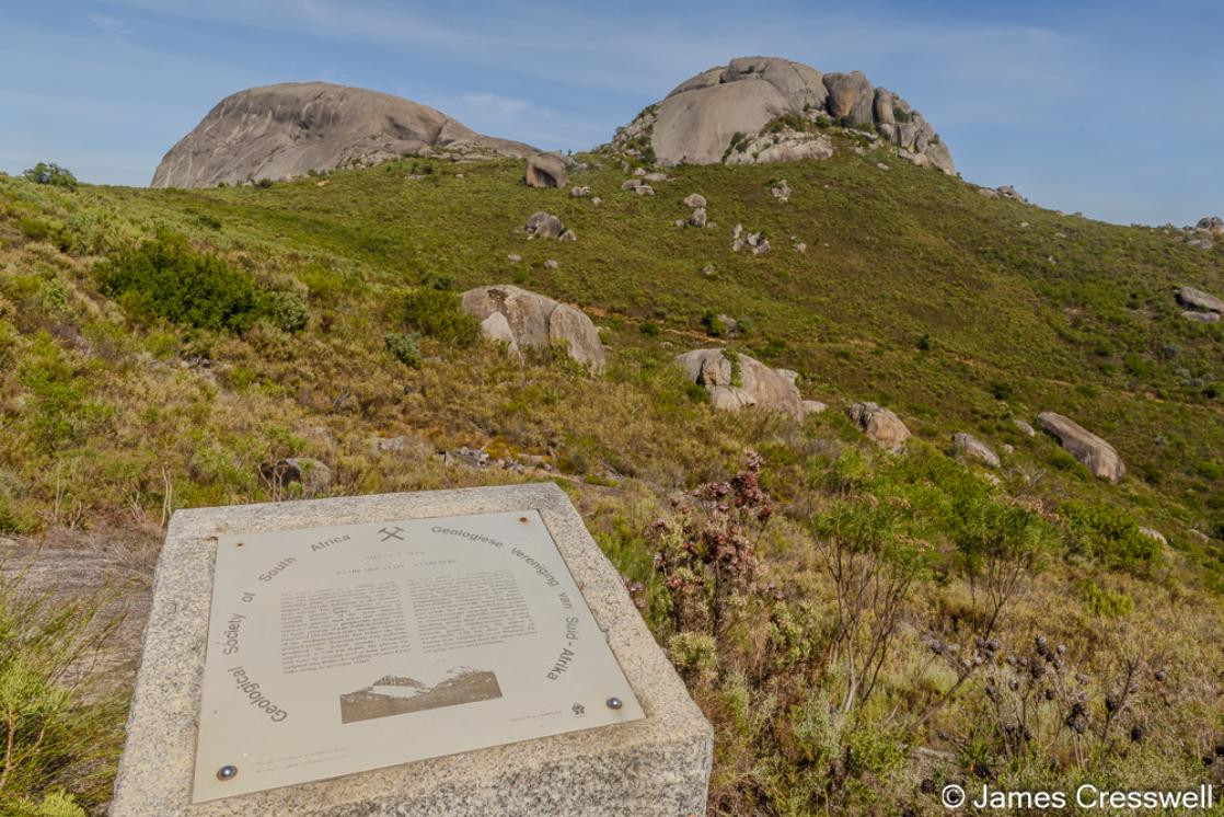 A photograph of the Geological Society of South Africa plaque on Paarl Rock with Bretagne and Gordon Rocks in the background. All the rocks are part of a Cambrian aged granite pluton.
