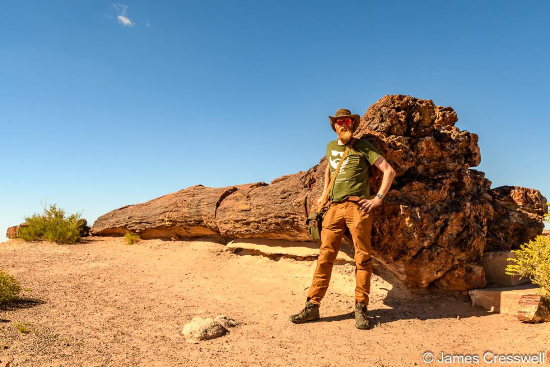 A photograph of James Cresswell of GeoWorld Travel with a fossilised tree trunk, Old Faithful at Petrified Forest National Park, USA